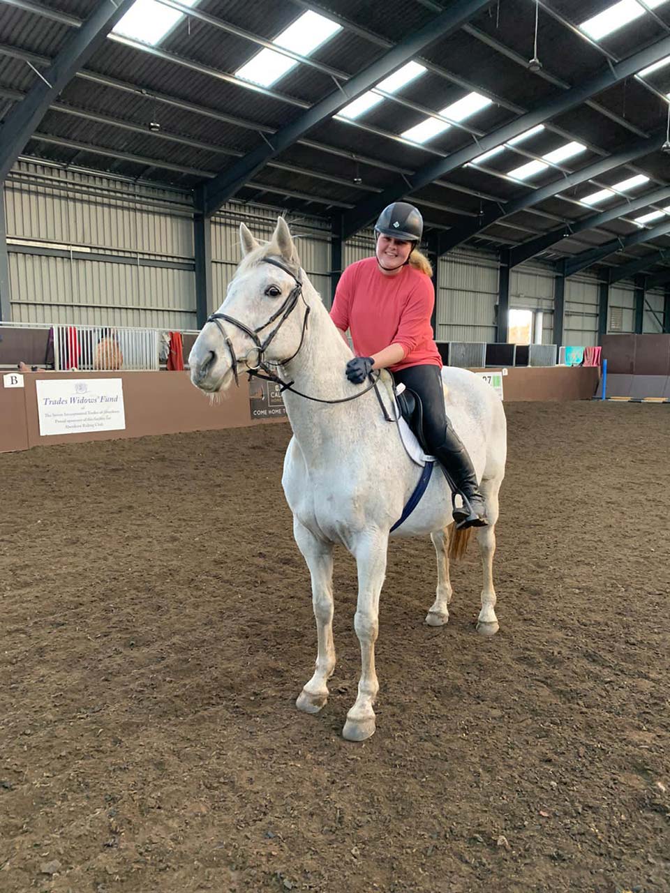 A person in a red shirt riding a gray horse in an indoor arena